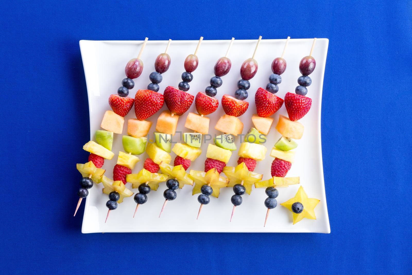 Healthy fruit kebabs with tropical seasonal summer fruit arranged diagonally on a modern rectangular plate over a texture blue background, viewed from above