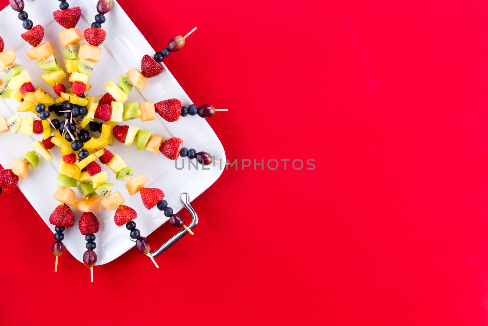 Festive arrangement of fresh fruit kebabs with exotic and tropical summer fruit arranged on a tray over a red party background with copy space, overhead view