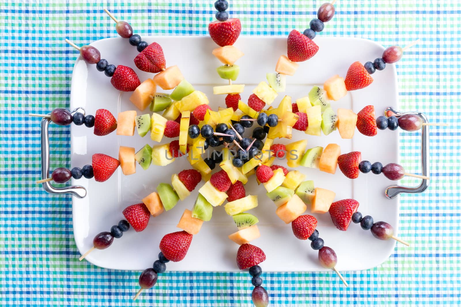 Presentation of colorful healthy fresh fruit kebabs on a summer picnic table made with seasonal exotic and tropical fruit for a gourmet dessert, overhead view