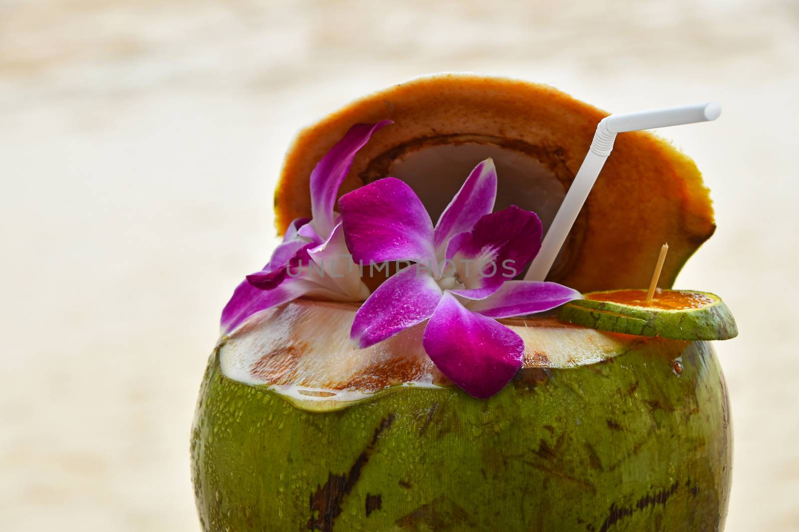Fresh open green coconut milk juice with straw decorated garnished with tropical purple orchid flowers and orange slice at sand beach background