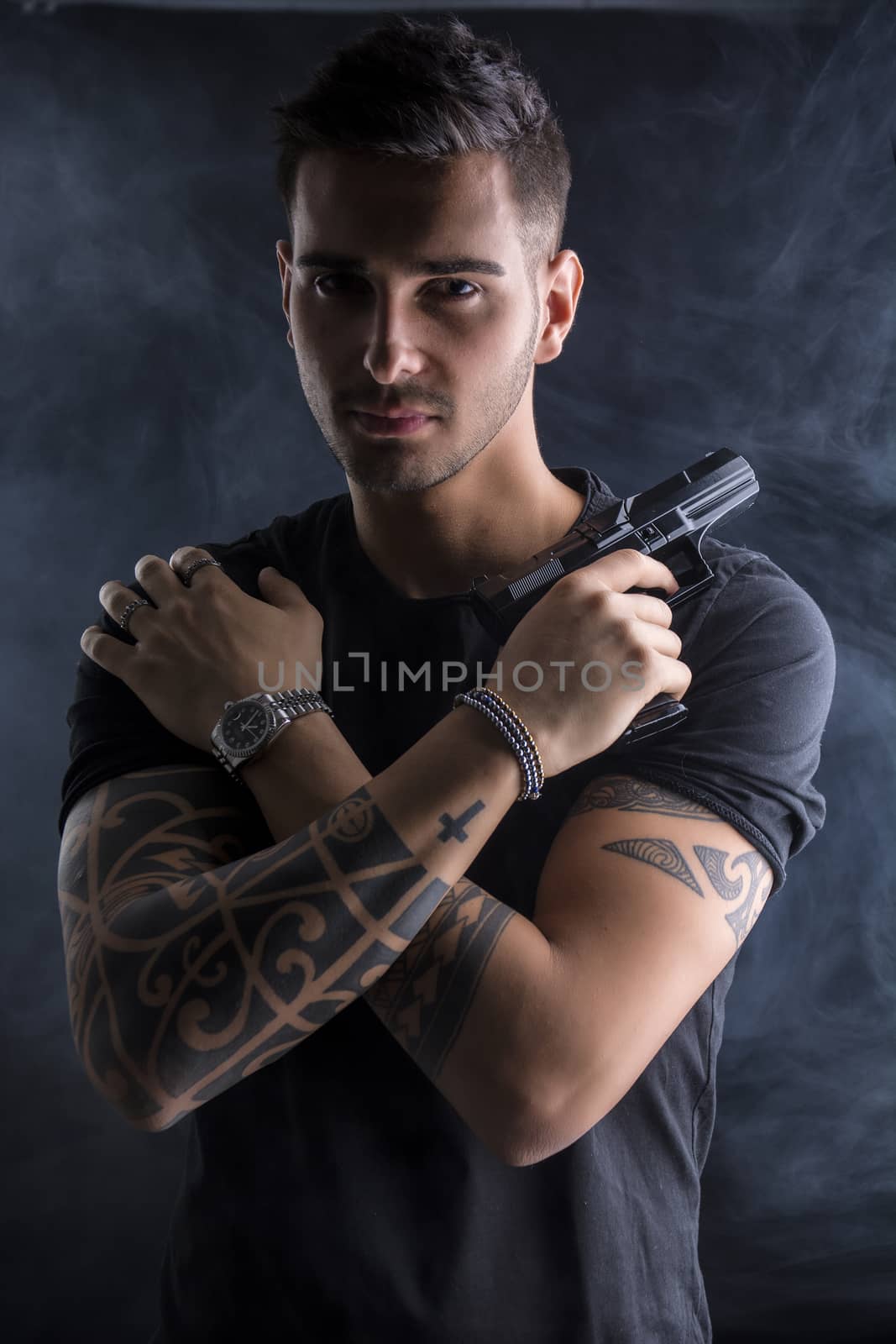 Young handsome man holding a hand gun, wearing black t-shirt, arms crossed on chest, on dark background in studio