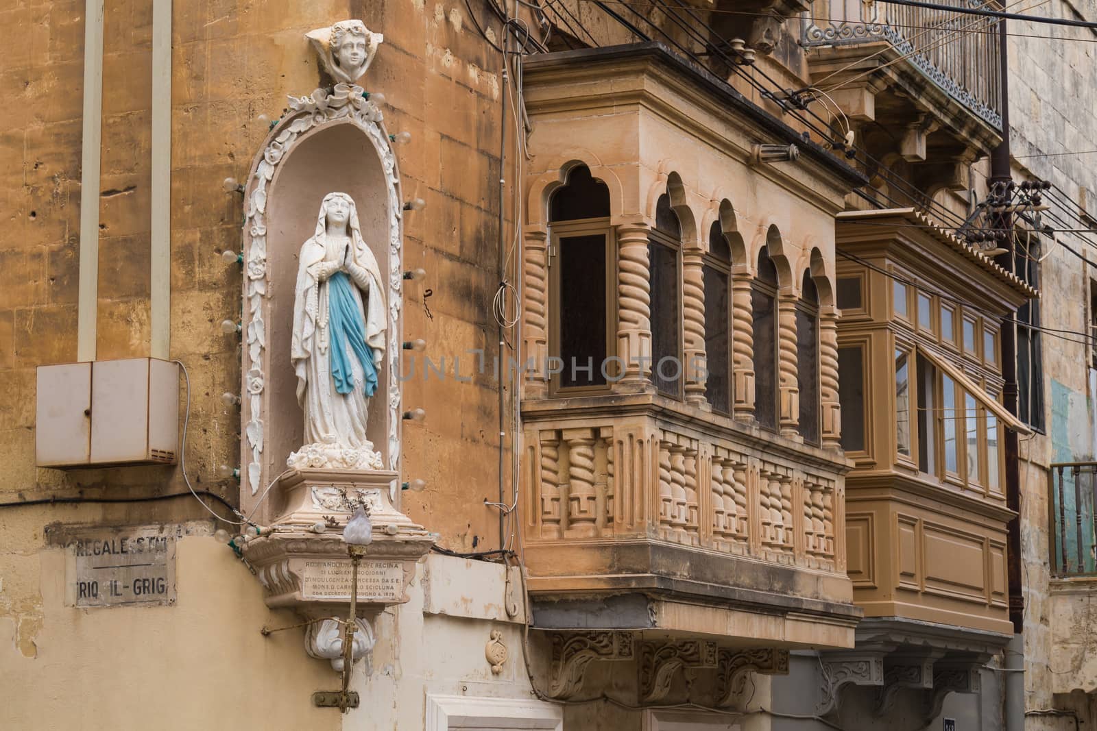 Ornate statue of a praying saint on the corner of a house with typical traditional balconies. Senglea, island Malta.