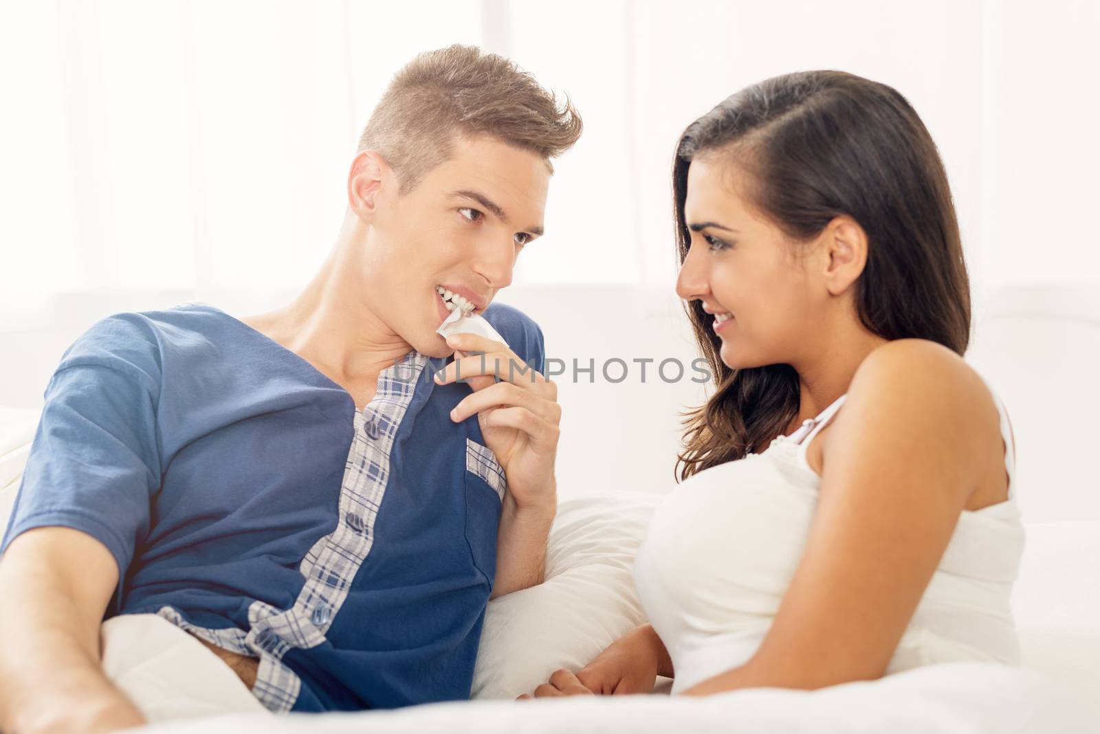 Young couple lying in bed looking at each other while a guy holding a condom in his mouth.