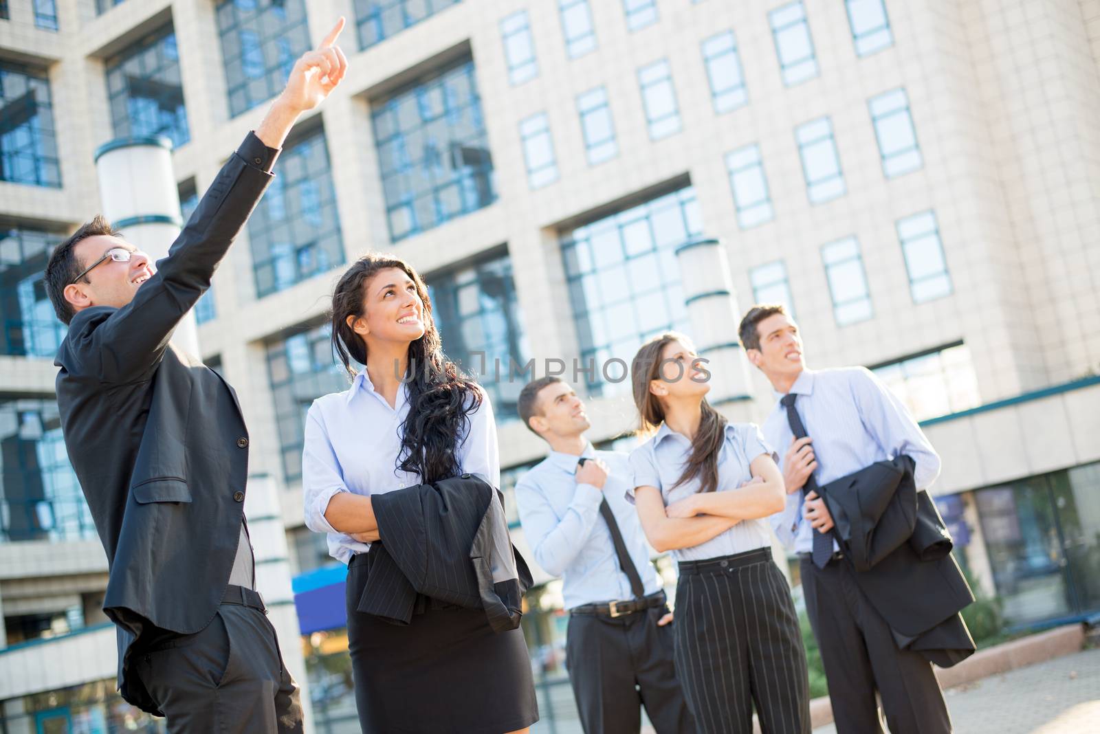 Elegantly dressed young businessman standing in front of large business building pointing his outstretched finger his colleague and his team to the top of the business complex where his office is located.