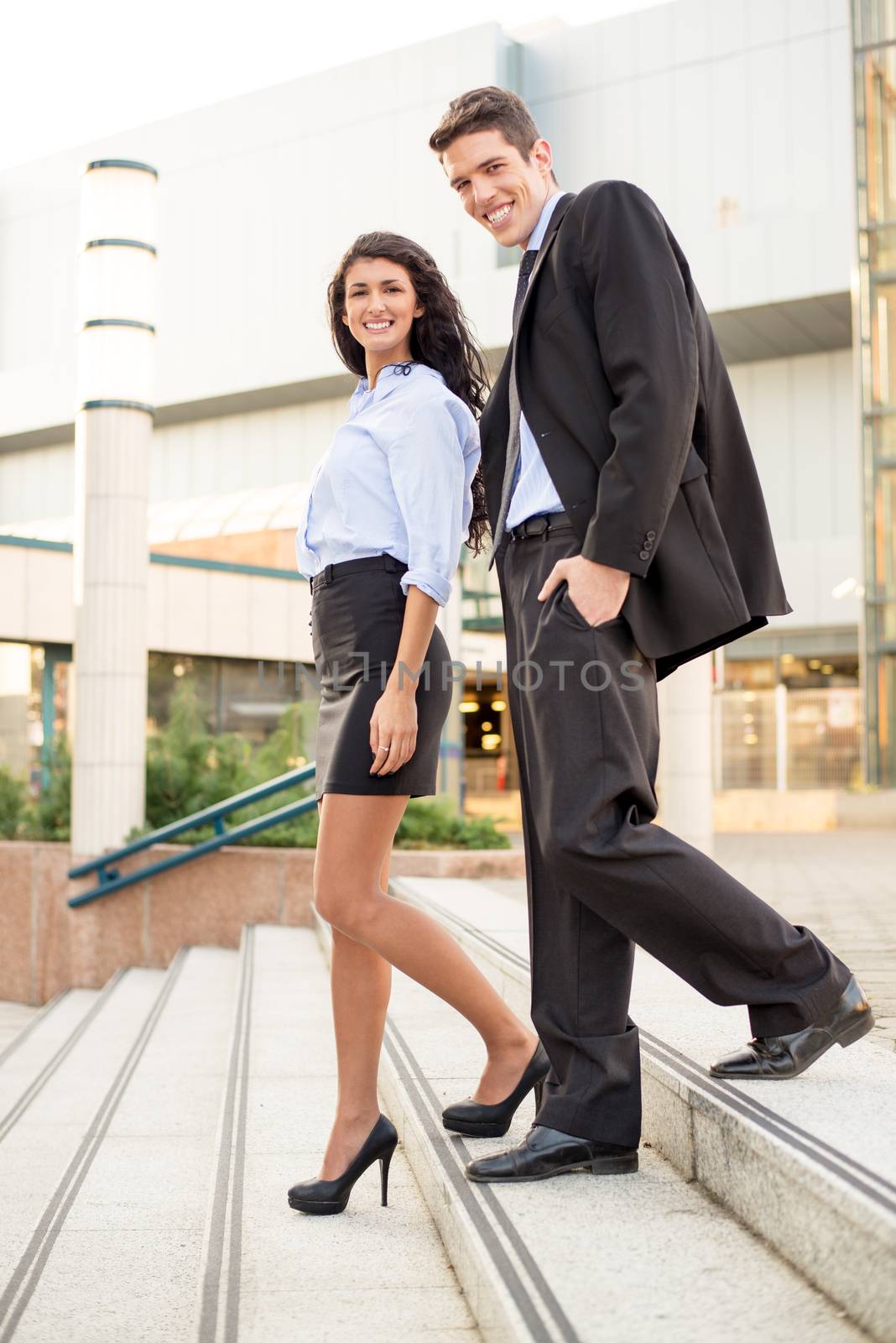 Young businessman and his colleague coming down the stairs of business building smiling looking at the camera.