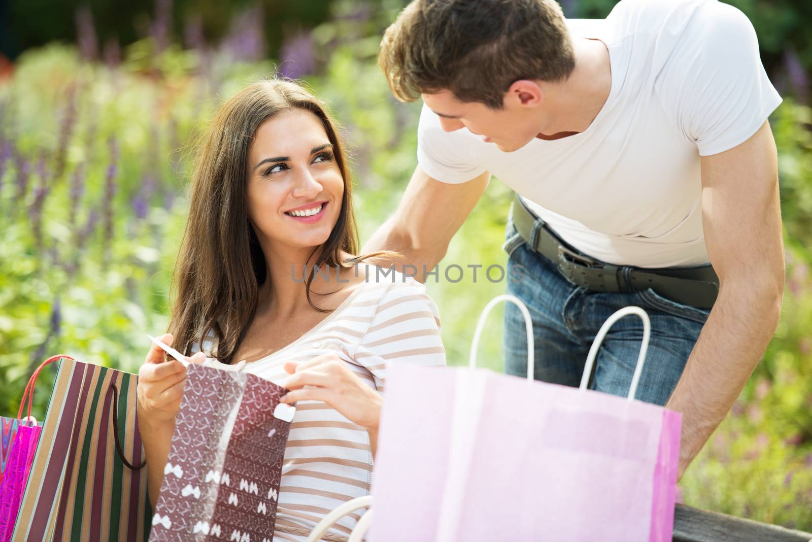 A young girl sitting on a park bench enjoying the beautiful sunny day, resting from shopping in addition to a bunch of shopping bags, holding a bag in his hand with a smile, looking at her boyfriend who was behind the bench leaning toward her face.