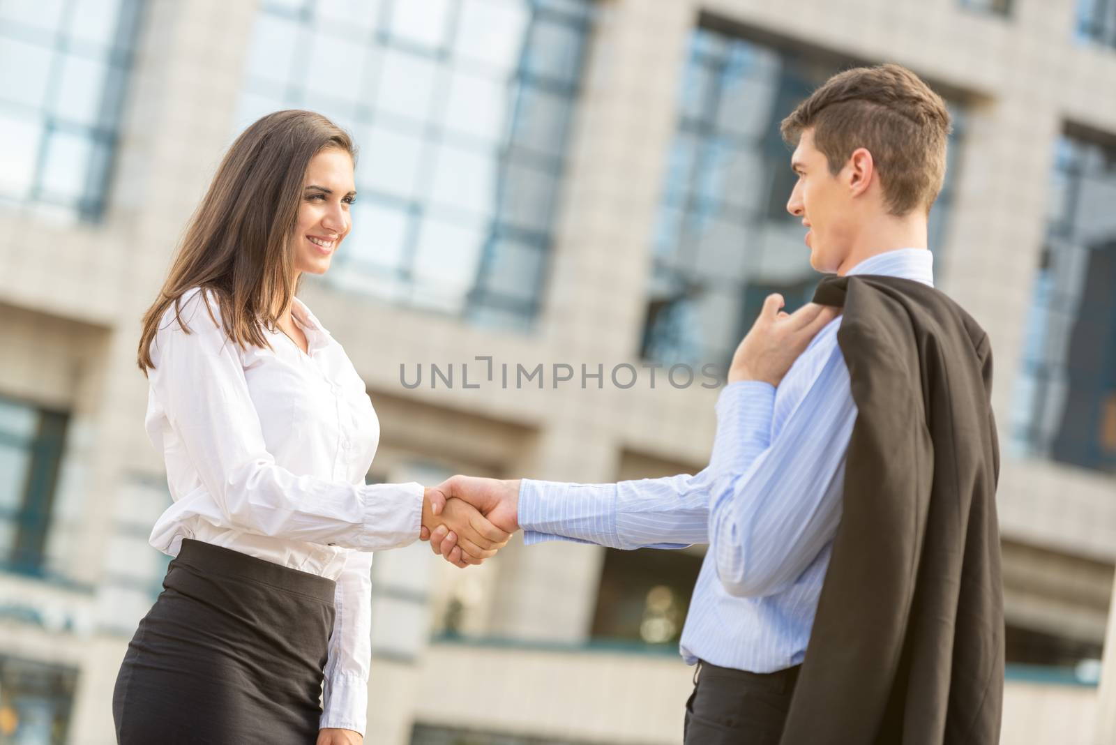 Young businessman and businesswoman, elegantly dressed, shaking hands in front of the company by looking at each other smiling happy business cooperation.