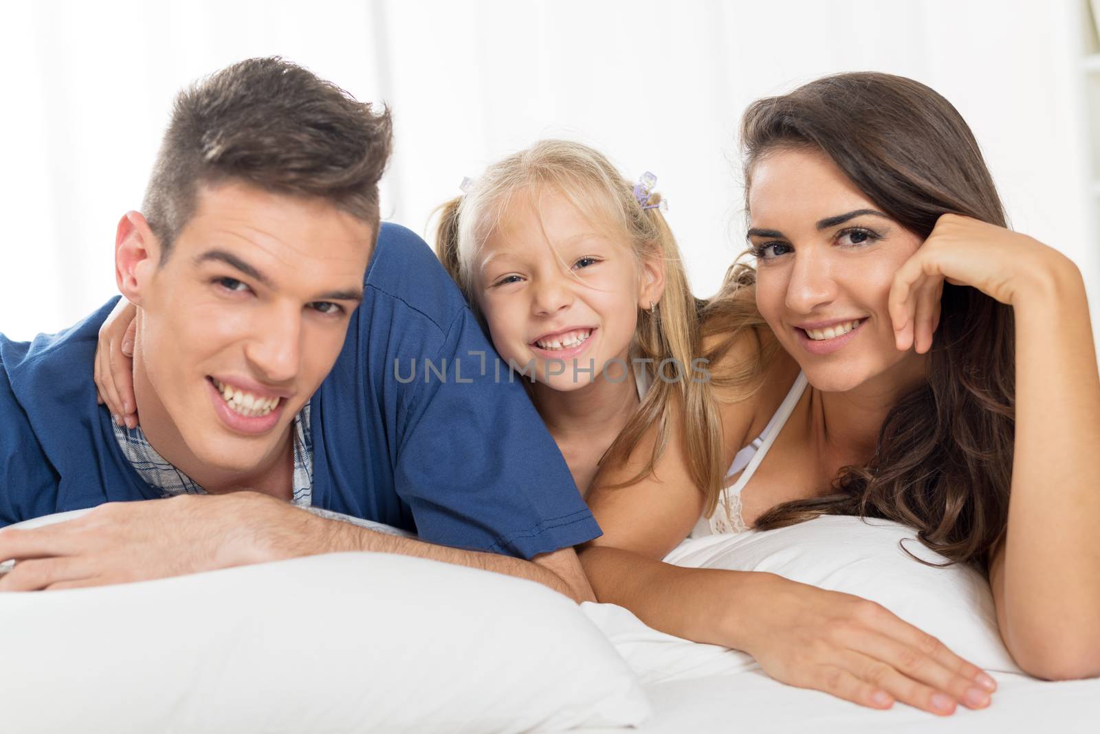 Young parents with their sweet daughter lying in bed looking at the camera with a smile.