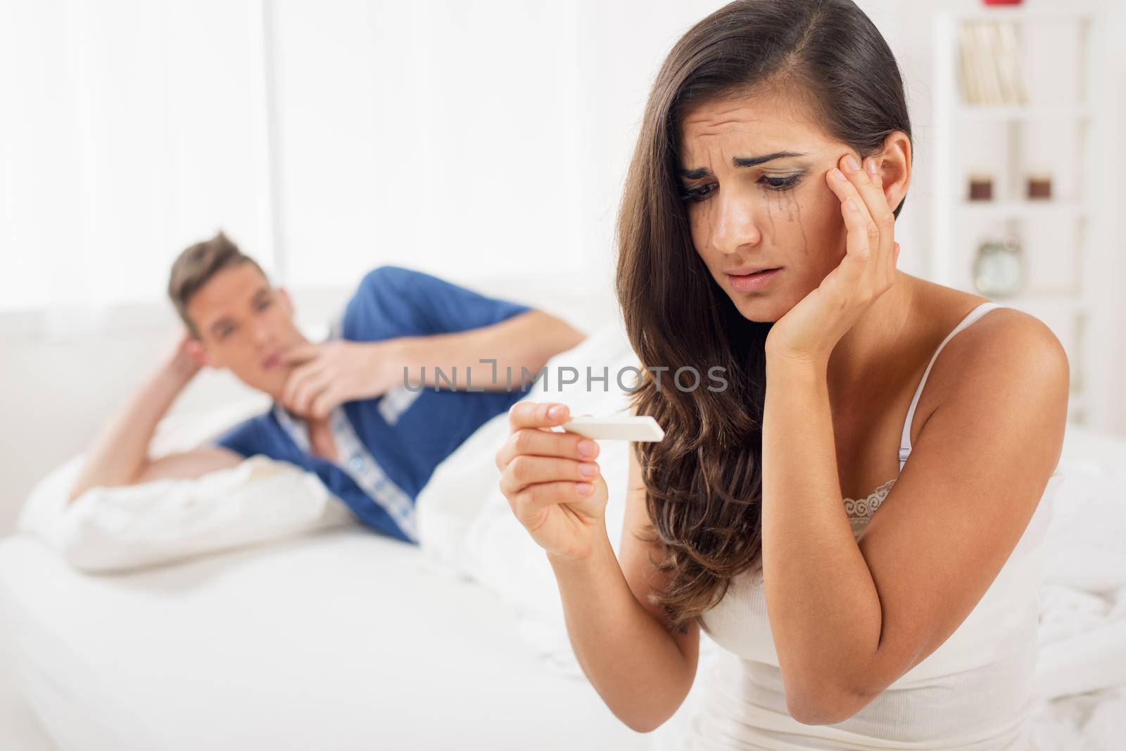 Young crying woman sitting on the bed and looking at a pregnancy test while in the background sees her partner lying in bed and looking at the woman.