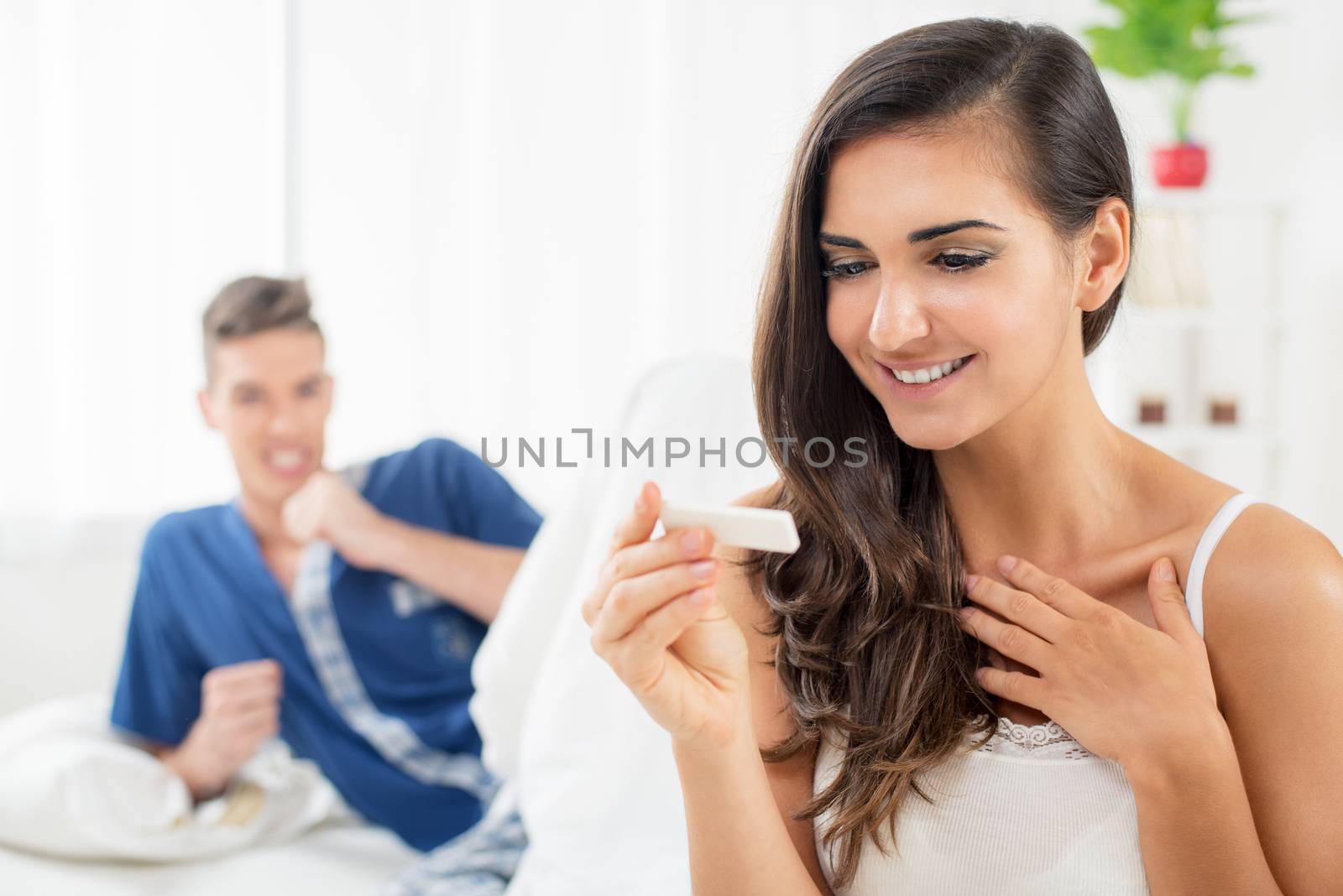 A young woman sits on a bed with a happy expression on her face looking at a pregnancy test, while in the background see her partner who is happy with the test results.