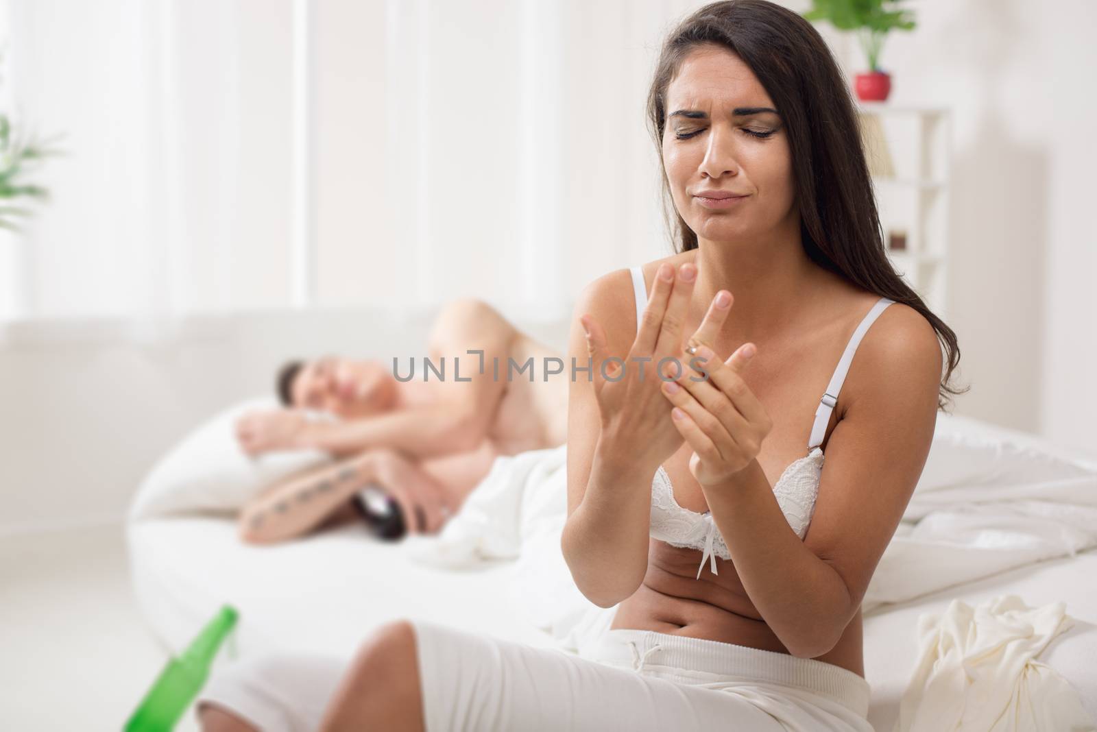 A young woman, just awakened, sitting on the bed with a sad expression on her face trying to take off the wedding ring from her finger, and next to her sleeping man hugging a bottle of drink.