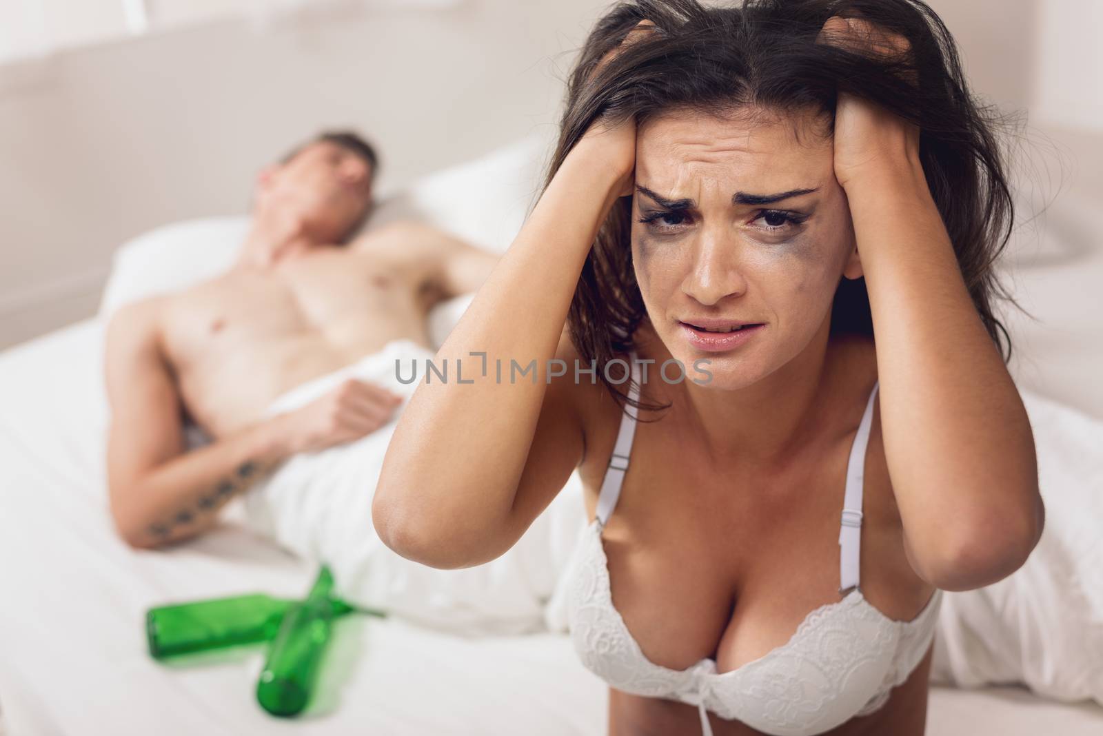 A young woman in a bra sits crying on the bed holding her head while next to her in bed sleeping next to her violent partner with empty bottles.