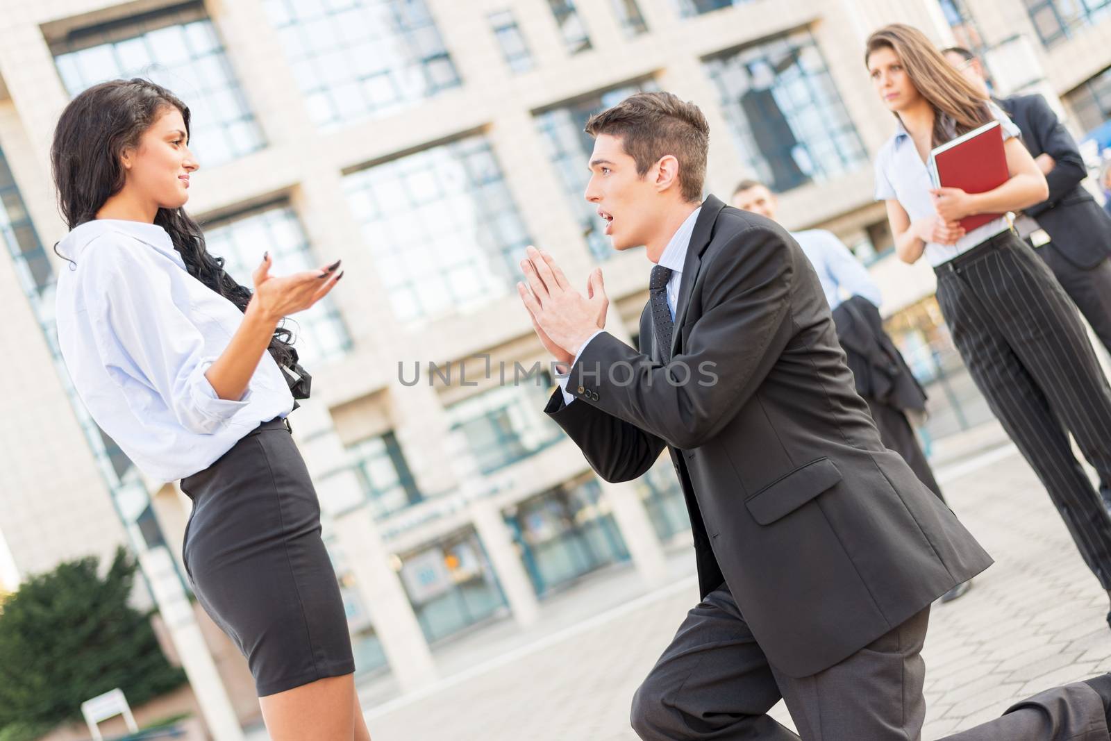 Young handsome, elegantly dressed businessman on the street in front of office building, kneeling with folded hands in front of a young attractive business woman while they watching colleagues.