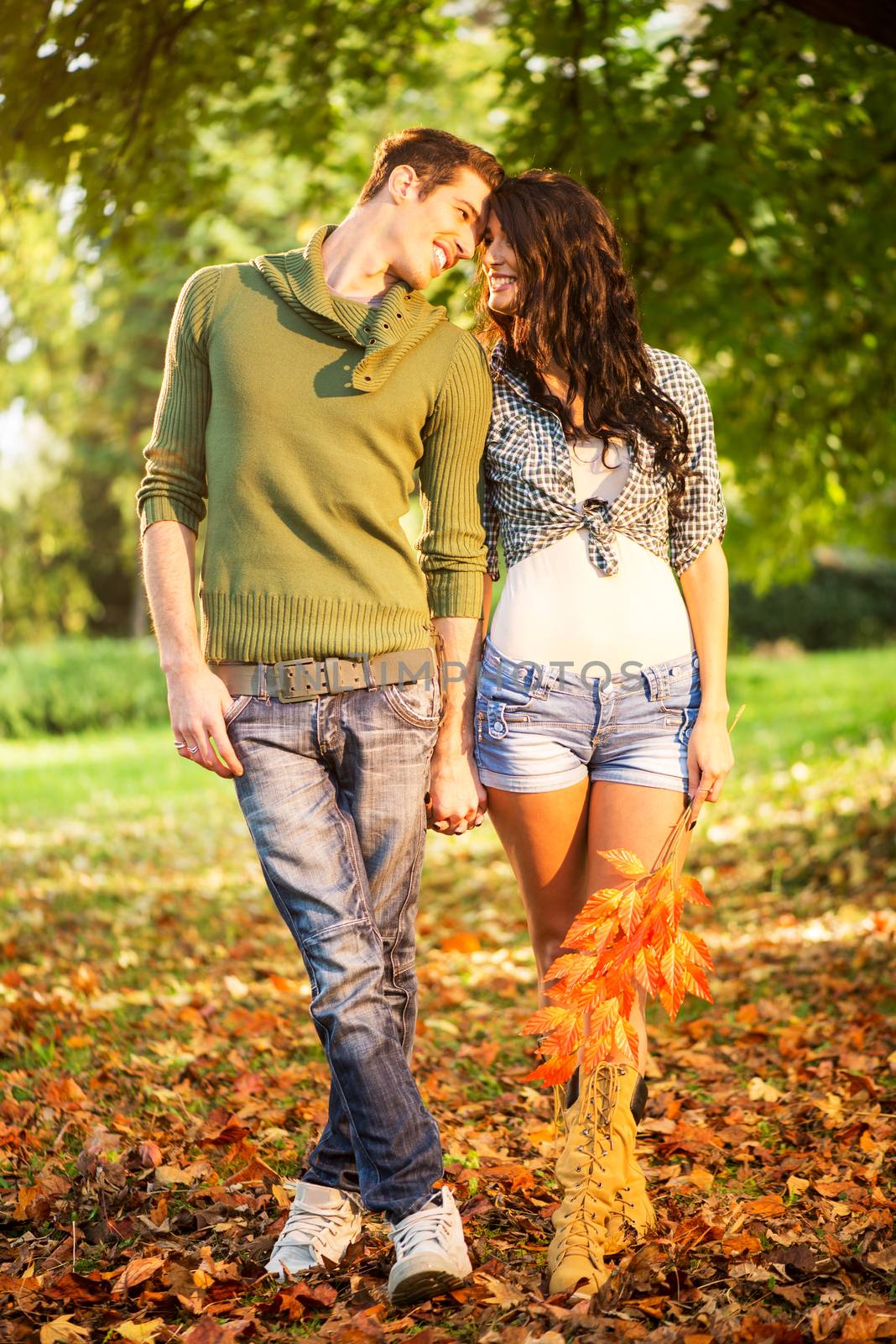 Young heterosexual couple in the park, walking on fallen leaves, holding hands and looking with a smile at each other.