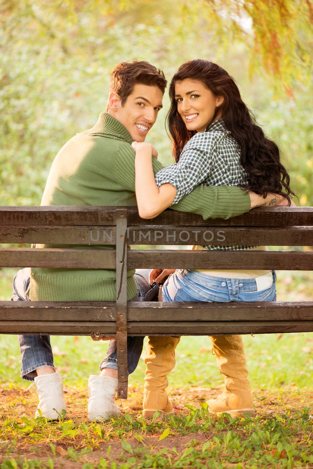 Photo of a young heterosexual couple hugging sitting on a park bench, behind their back looking at the camera with a smile on their faces.