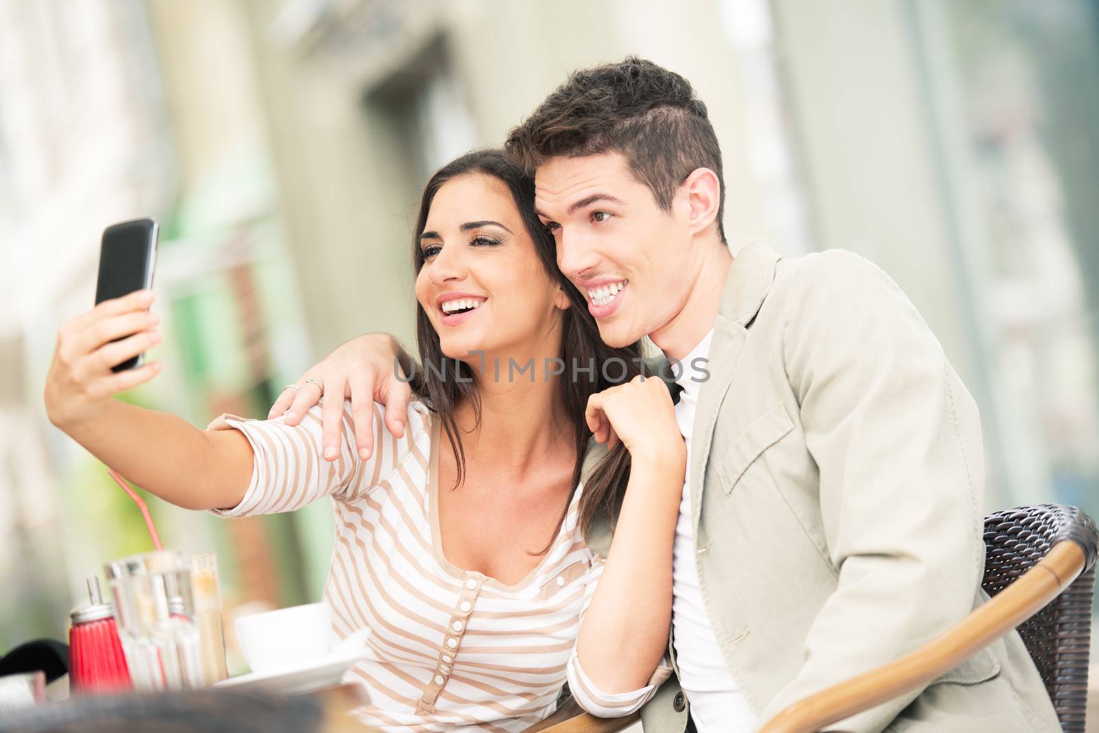 Young heterosexual couple sat embraced in a cafe, and a girl with her cell phone photographs of herself and her boyfriend.