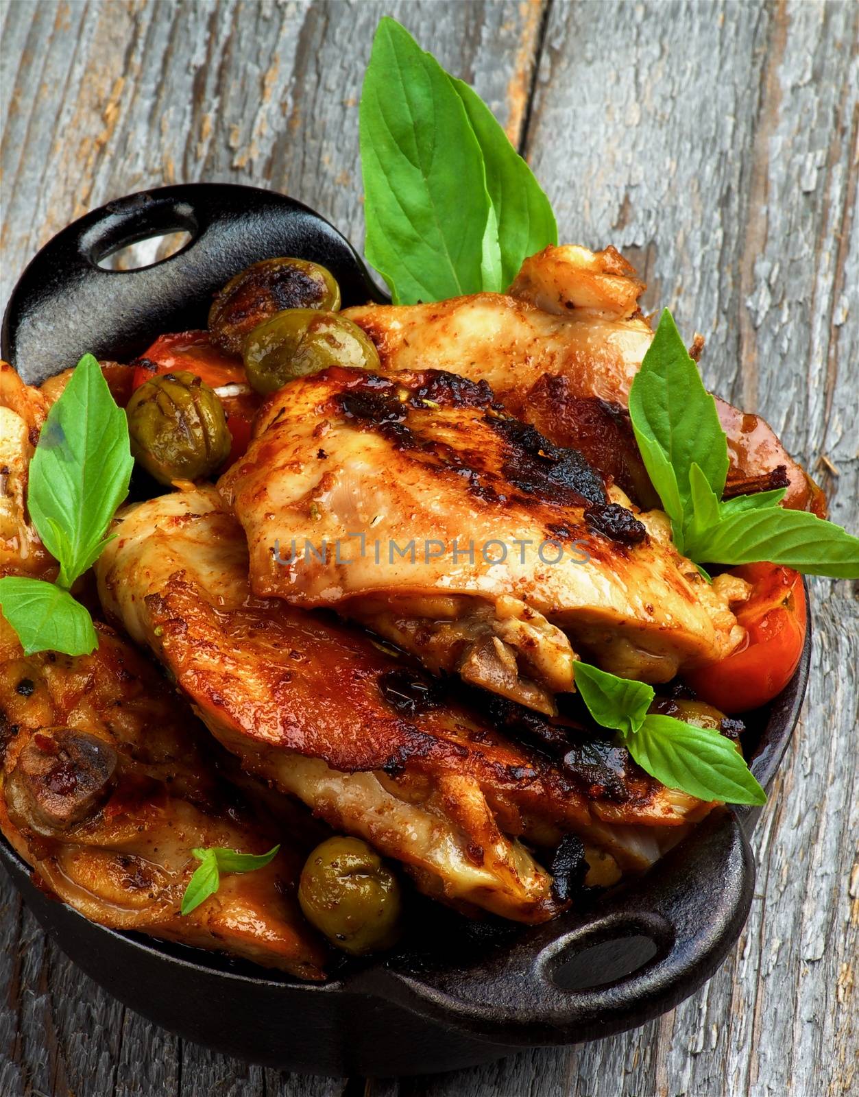 Delicious Roasted Chicken Thighs with Green Olives, Cherry Tomatoes and Basil in Black Fry Pan closeup on Rustic Wooden background