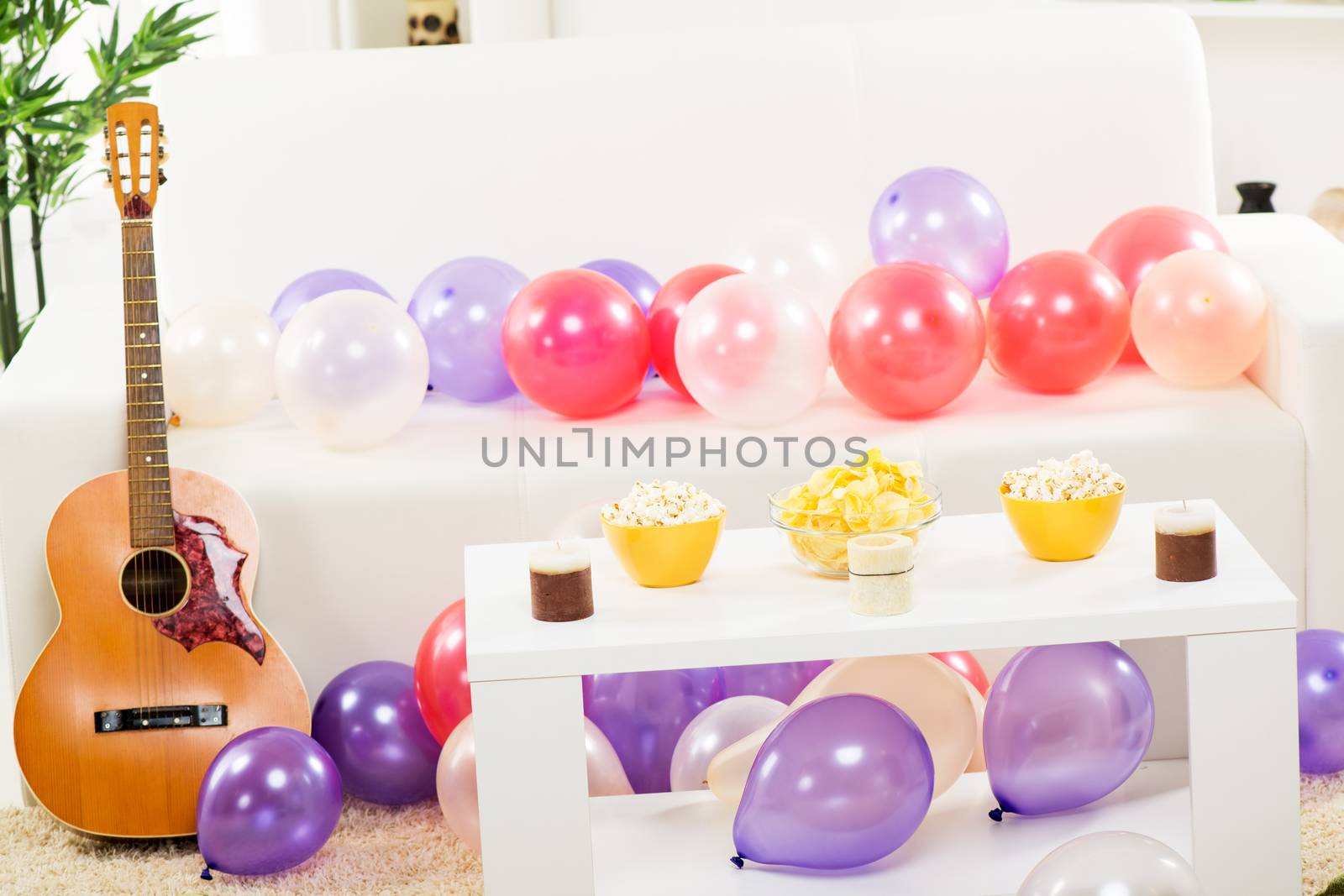 Room with a couch where is leaned acoustic guitar, couch and floor are covered with balloons, and on the table in front of the couch, there are decorative candles and snacks.