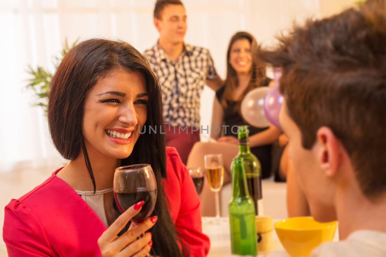 A young pretty girl with a glass of wine in hand, with a smile looking at the guy in front of her, while in the background see another young couple in the ambient house parties.