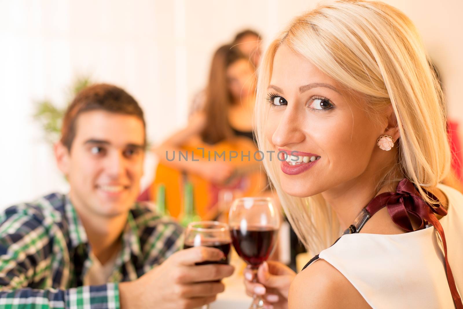 The young beautiful girl and young guy knocking the glasses toasting with wine while in the background in the ambient house parties see the girl who plays acoustic guitar.