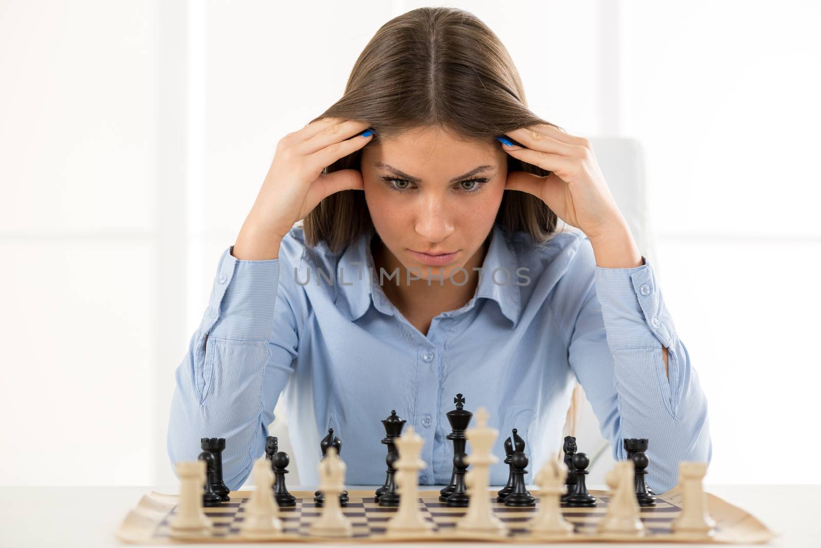 A young businesswoman is sitting in front of a chess board and thinking staring at the figure, holding hands on her head.