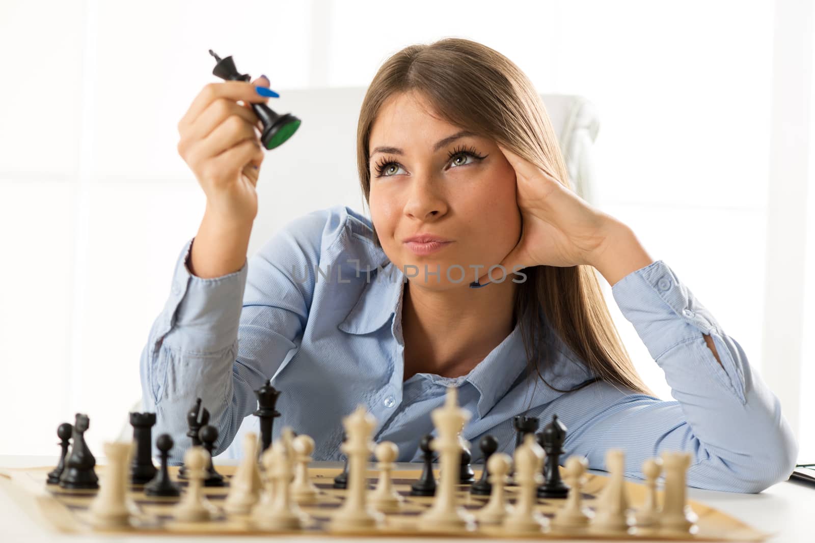 Young pretty businesswoman sitting at the table over a chessboard resting on her hand looking at the black king chess piece that holds in the other hand.