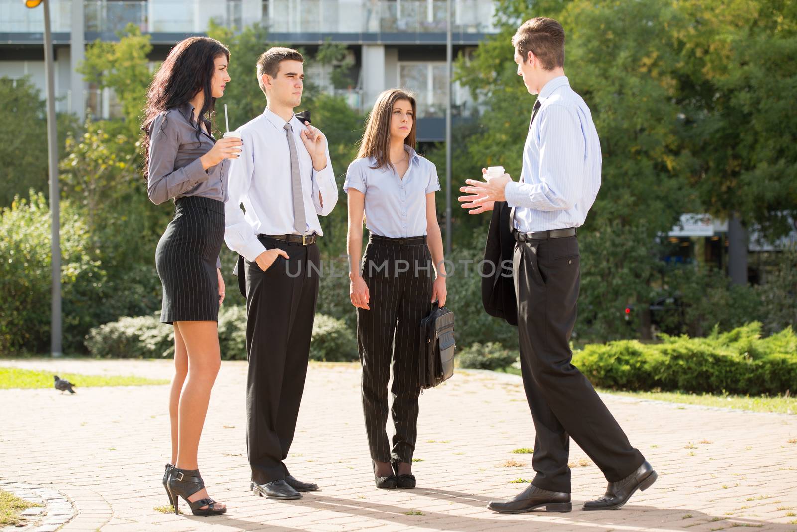 Group of young business people standing in a park near their company on the coffee break, talking to each other, enjoying the beautiful day.