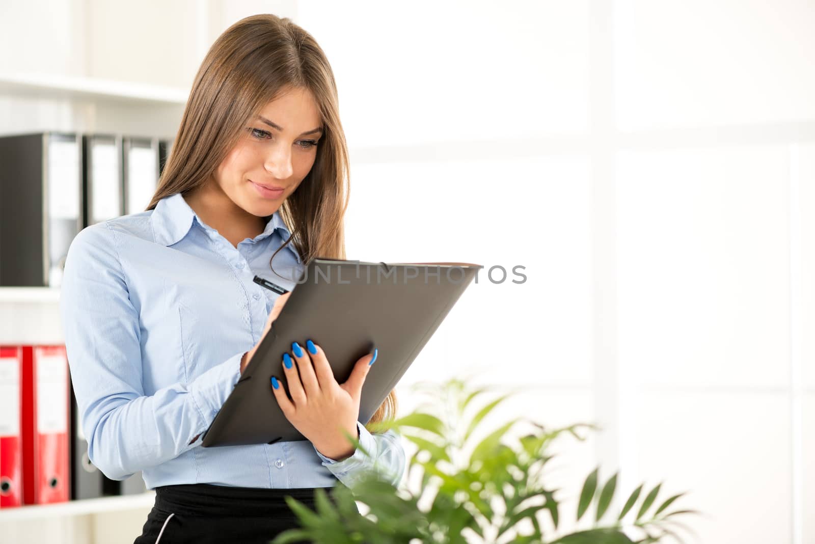 Young businesswoman is standing in an office and write on the folder that holds in her hands, in the background you can see the shelves with binders.