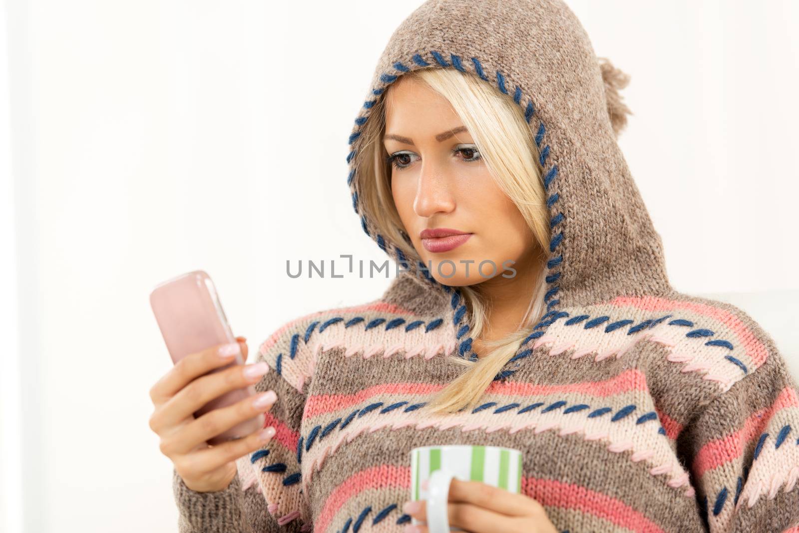 Beautiful young woman in a wool sweater with a hood on her head, looking at mobile phone that holds in her hand, and in the other hand holding a cup.