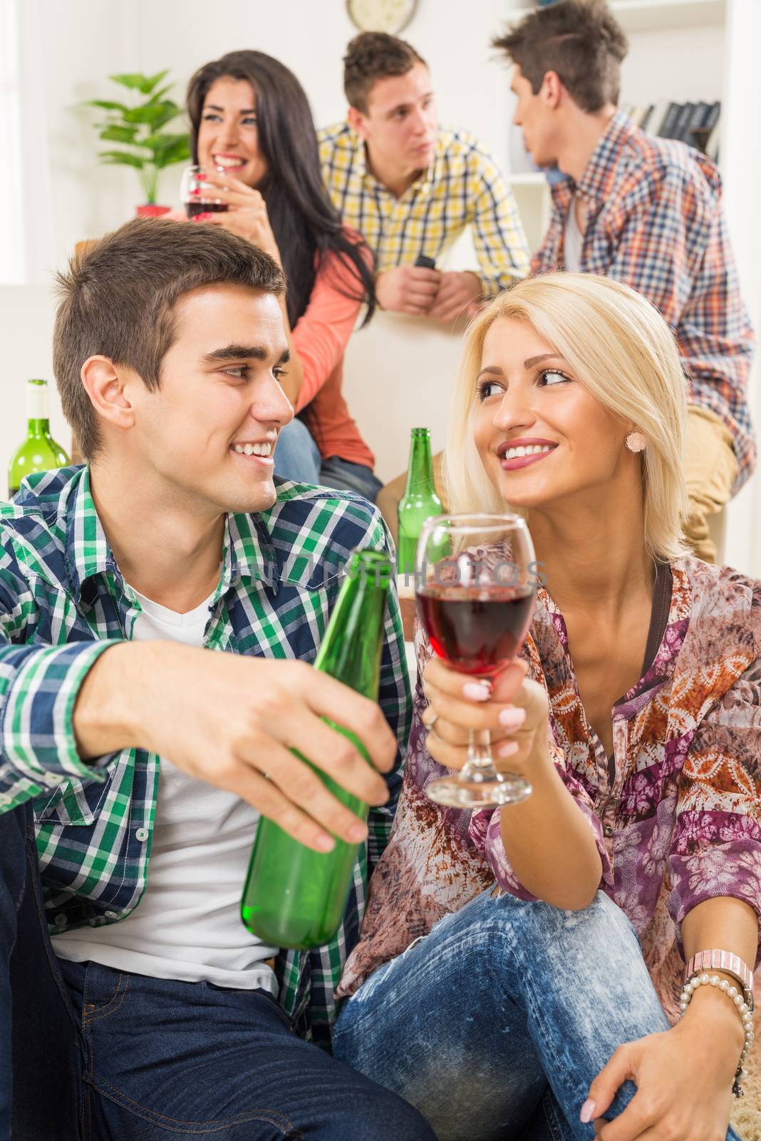 Young couple at a home party, sit on the floor and knocking with drinks, in the background you can see their friends who are sitting on the couch.