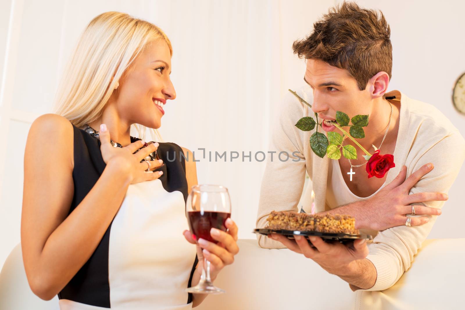 A young man with a rose in his mouth gives beautiful blond girl birthday cake, pleasantly surprised girl looking at him with a smile on her face.