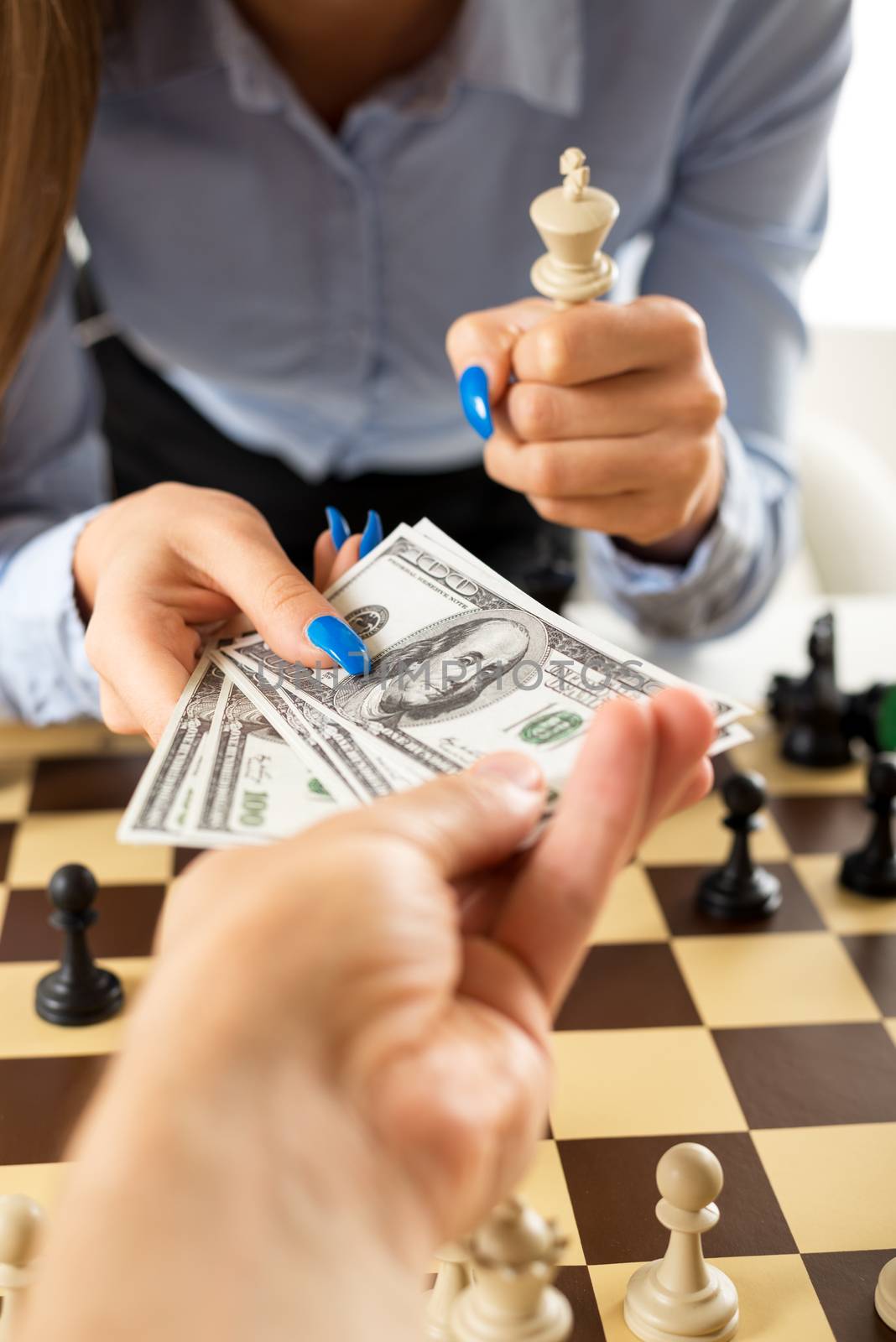 Close-up of female hands with painted fingernails over the chessboard. One hand takes money from the man's hand, and the other female hand holding in the hand white king chess piece.