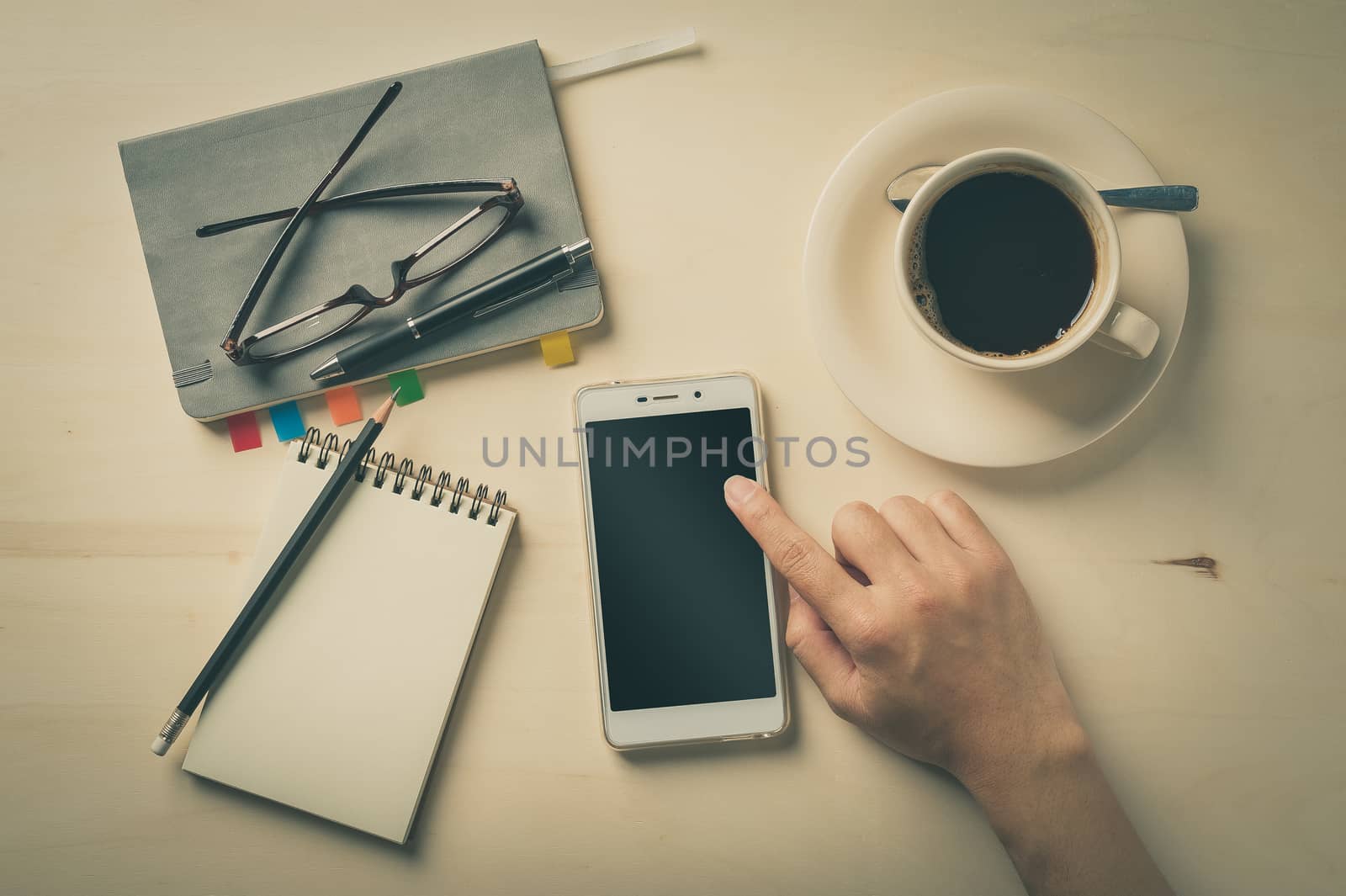 Male finger touching on smartphone screen with notebooks, pen, and coffee cup beside on wood table in morning time with vintage filter effect