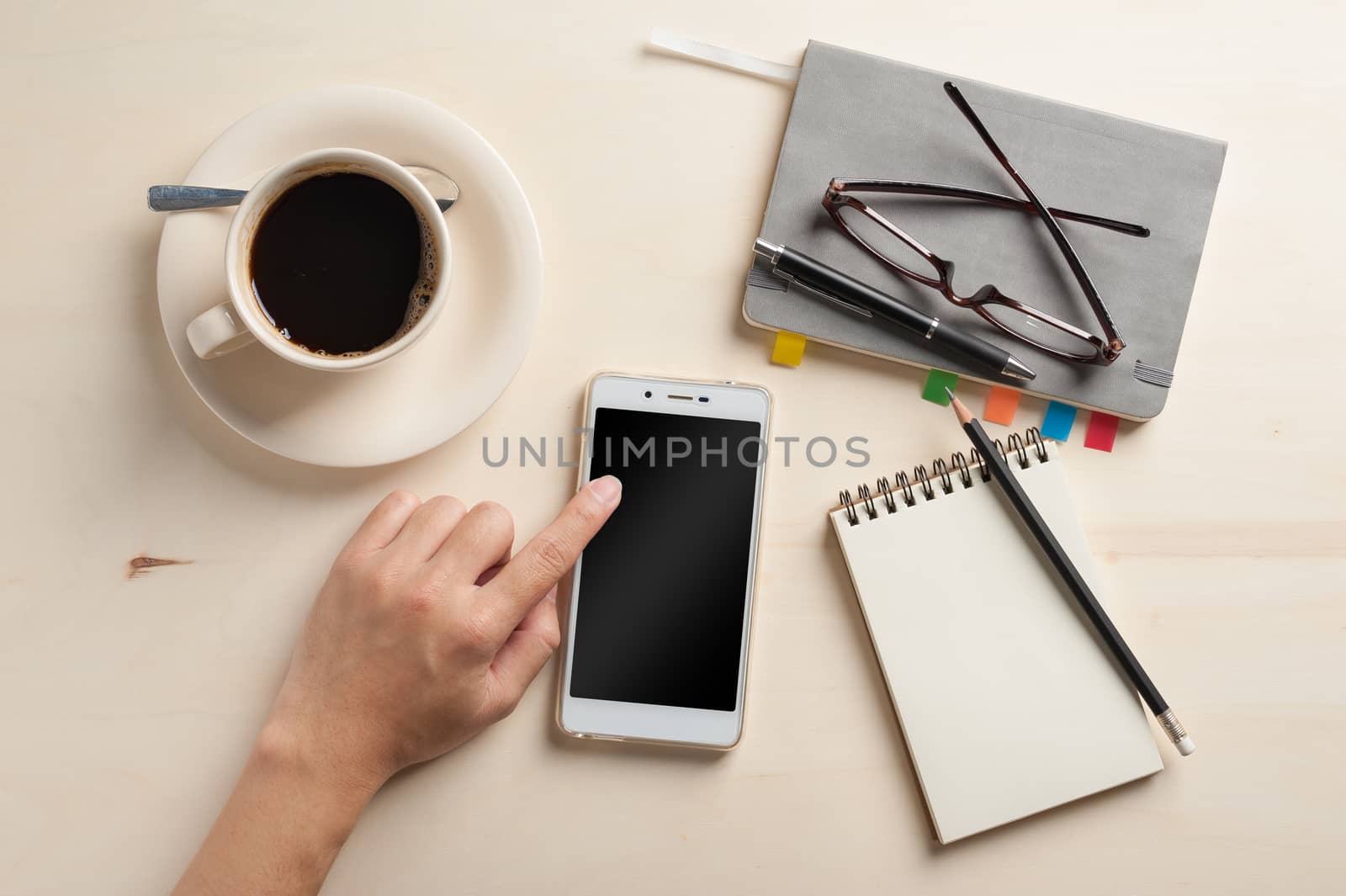 Male finger touching on smartphone screen with notebooks, pen, and coffee cup beside on wood table in morning time