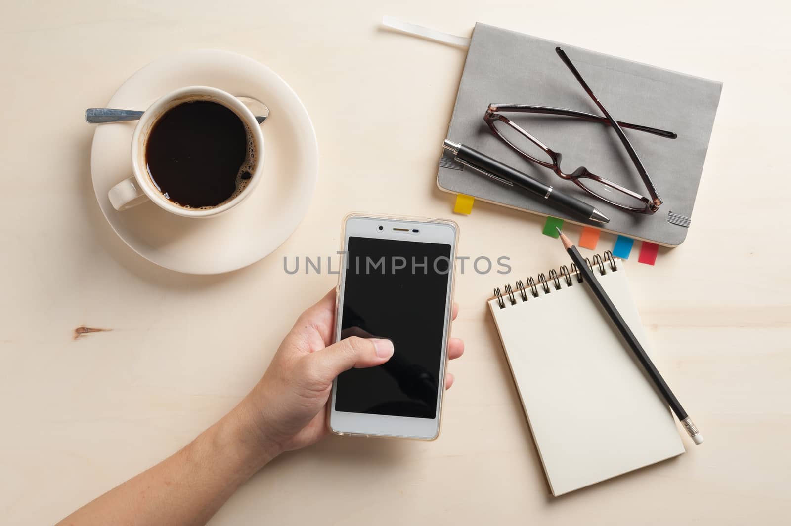 Male finger touching on smartphone screen with notebooks, pen, and coffee cup beside on wood table in morning time