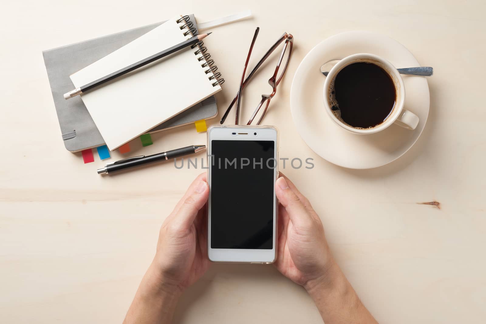 Male hand holding smartphone with notebooks, pen, and coffee cup beside on wood table in morning time