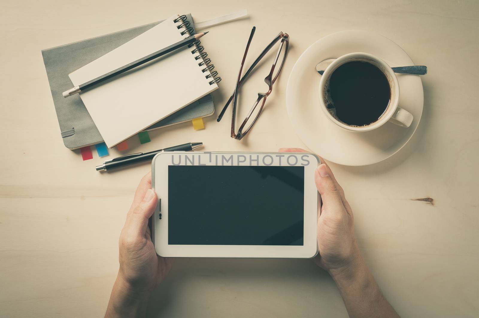 Male hand holding small tablet pc with notebooks, pen, and coffee cup beside on wood table in morning time with vintage filter effect