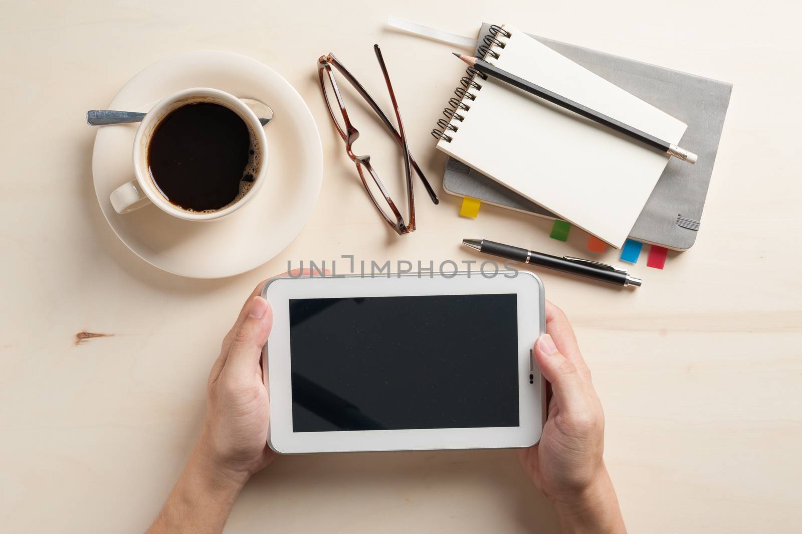 Male hand holding small tablet pc with notebooks, pen, and coffee cup beside on wood table in morning time