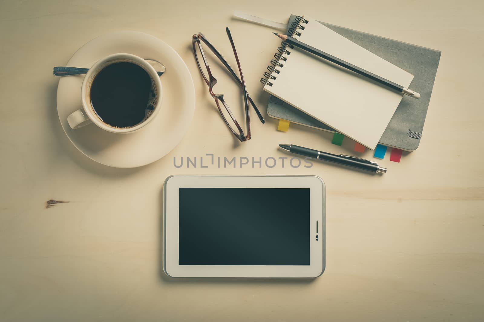 Small tablet pc, notebooks, pen, glasses, and coffee cup on wood table in cafe at morning time with vintage filter effect