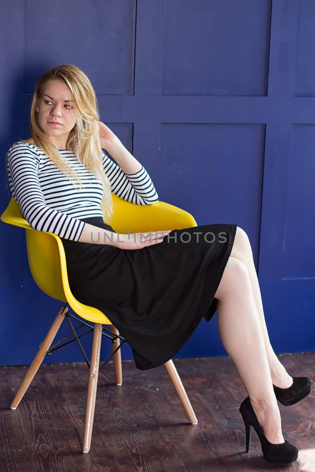 Blond girl in a long skirt and a vest sits on a chair next to the blue walls
