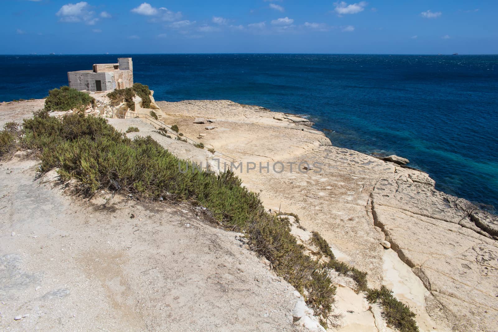 Sand color of the rocks on the coast of Mediterranean sea, city Marsaskala,island Malta. Building on the top of the hill. Bright blue sky, few white clouds.