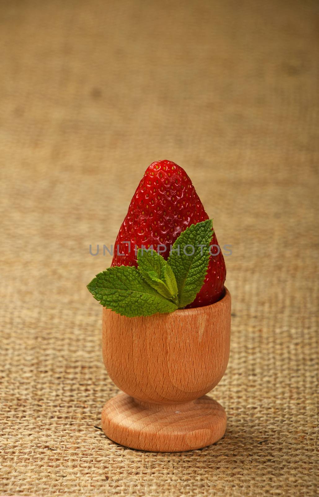 One big red mellow strawberry and green fresh mint leaves in wooden eggcup holder on natural burlap jute canvas background