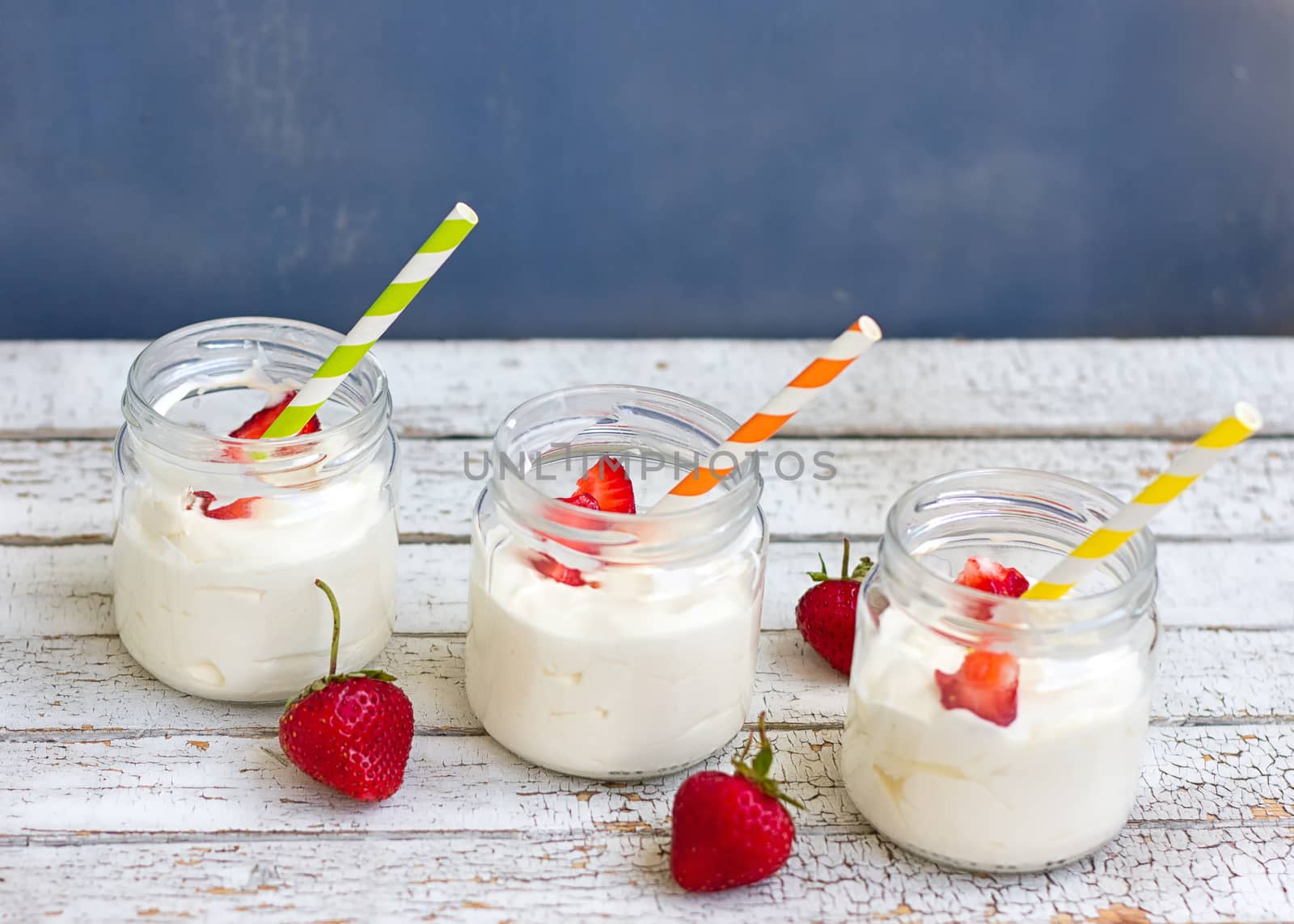 Three jars of yogurt with strawberries and colored tubes on a wooden board