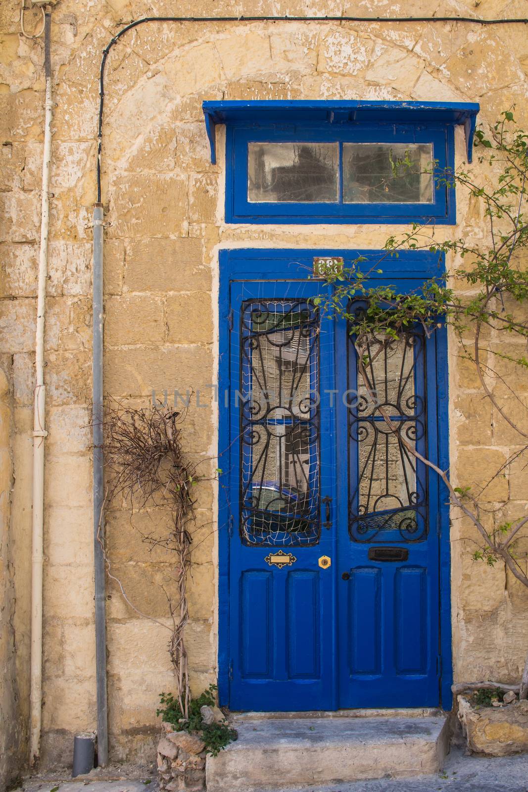 Wall of a stone building in the capital of Malta - Valletta. Bright blue wooden gate with two glasses with a reflection.