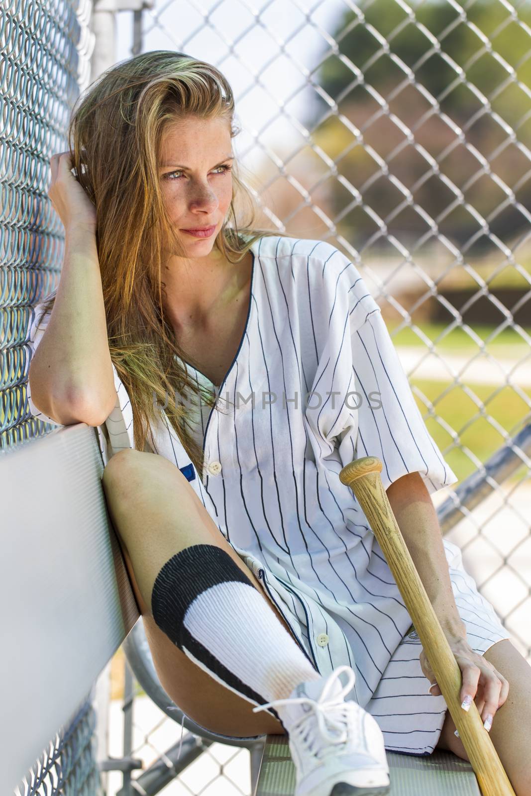 Blonde female model posing outdoors at a baseball diamond wearing baseball equipment.