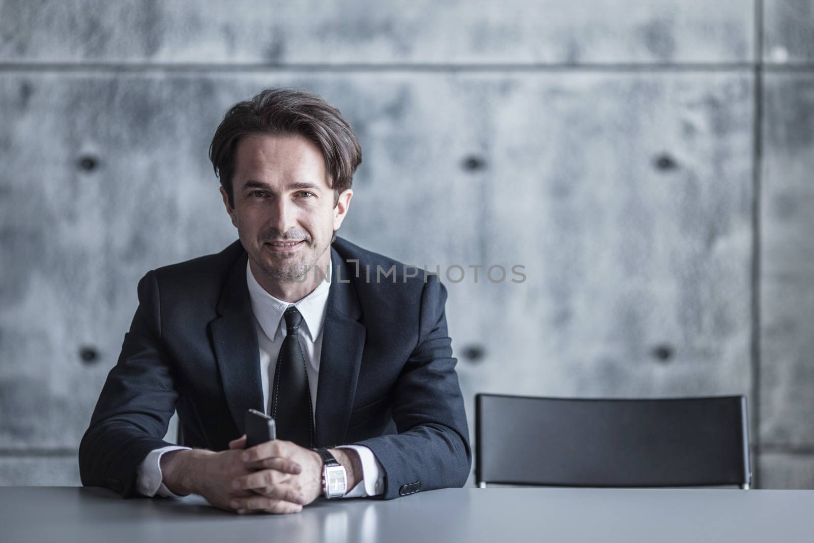 Portrait of a businessman with smartphone sitting behind the table over concrete wall background