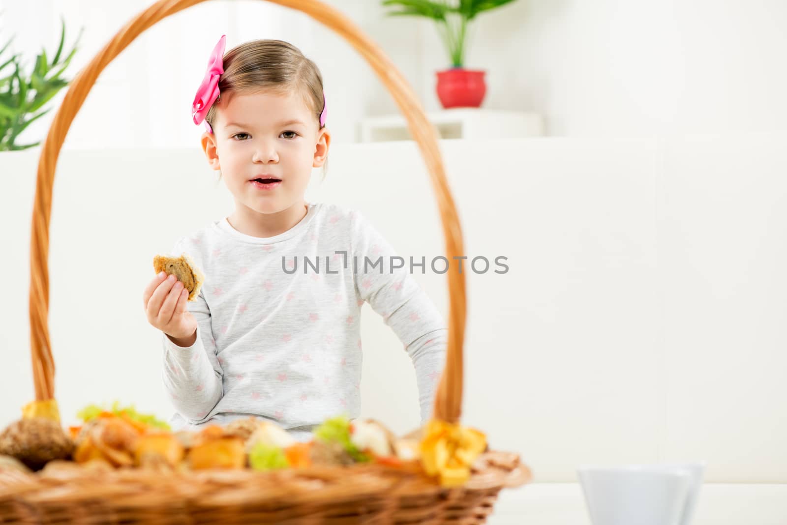 Portrait of cute little girl with hairclip, eating pastry, front wicker basket filled with tasty baked goods.