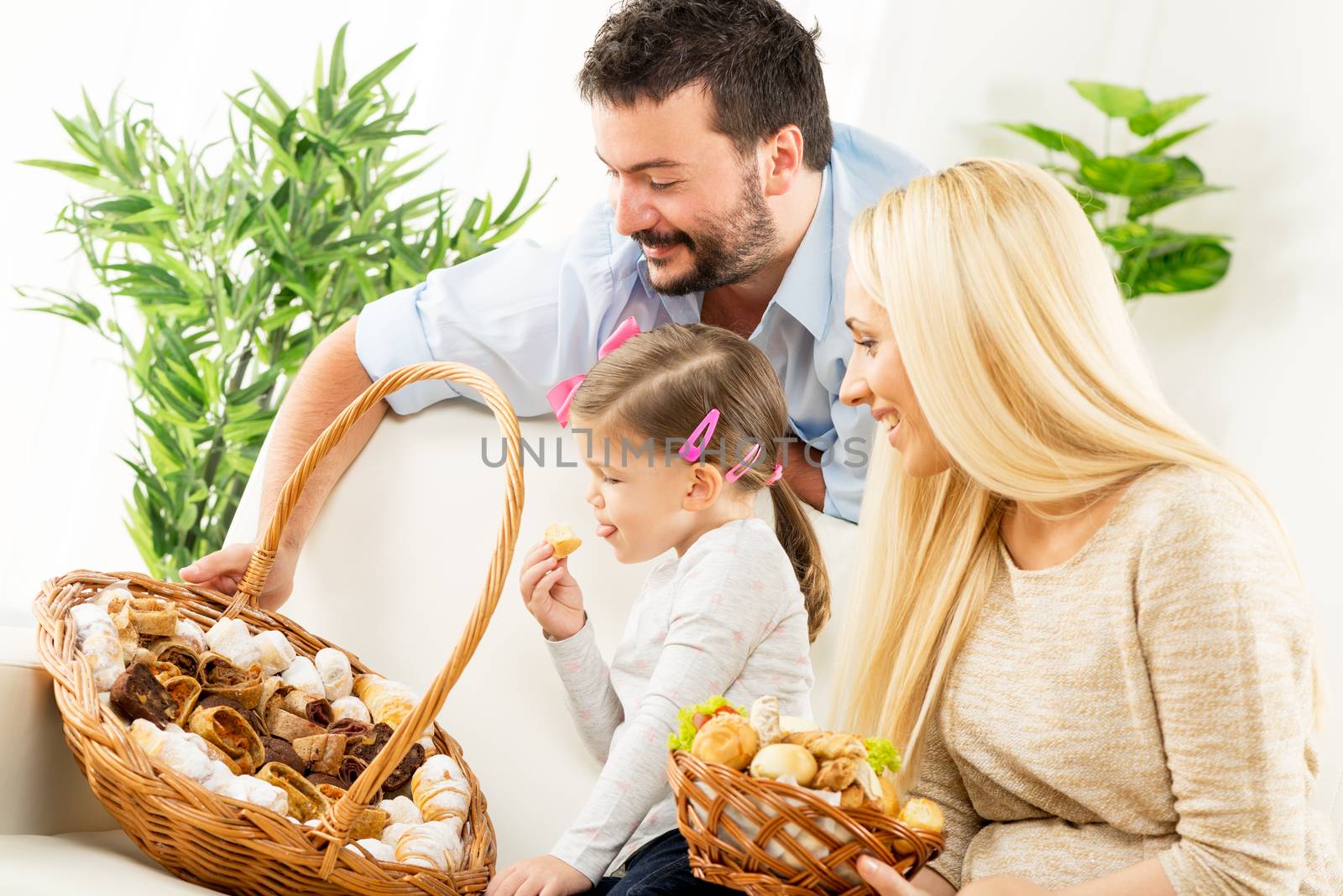 Happy family together, parents with daughter sitting on the couch in the living room in front of woven baskets filled with baked products, choosing the next snack.