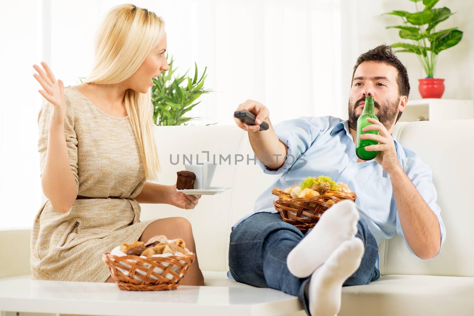 Young man sitting on a couch with his feet on the table, holding a bottle of beer in one hand, remote control in the other hand, in the knee had baskets with pastries, while sitting next to him pretty blonde woman shouting at him.