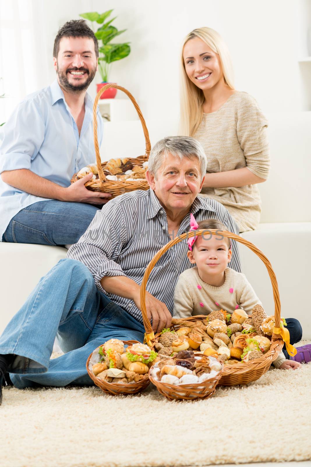 Senior man and a little girl are sitting on the living room floor. In front of them is woven basket with pastries, and behind them young parents are sitting on the couch and hold a basket of pastries.